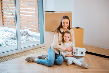 happy mother hugging cute daughter while sitting on floor in new home