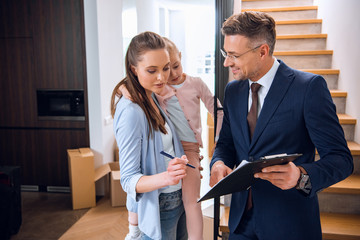 attractive woman holding in arms daughter and signing document near handsome broker holding clipboard