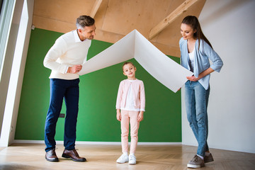 cheerful daughter standing under paper roof in hands of happy parents