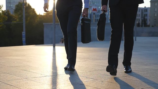 Feet of two businessmen with briefcases walking in city street at sunset time. Partners commute to work together. Confident guys being on his way to office. Colleagues going outdoor. Slow motion
