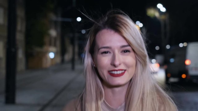 Outdoor portrait of young woman with long blond hair, red lipstick, and stylish look in the evening city turns to camera and smiles. Night city life. NY. New York