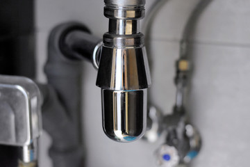 Chrome sanitary elements and pipes with selective focus under bathroom sink. Sewerage system with steel fitting connector elements. Home water plumbing construction with metal siphon under basin.