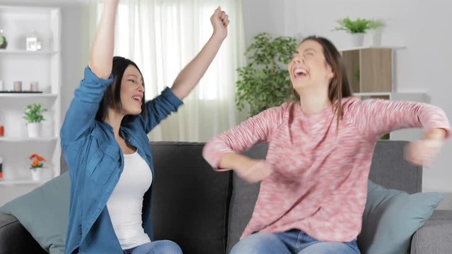 Two excited friends celebrating good news sitting on a couch in the living room at home