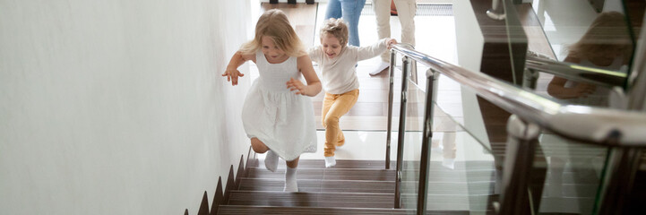 Happy kids go upstairs to second floor their new modern house, family moving day relocation and loan, buy first home concept. Horizontal photo banner for website header design with copy space for text - obrazy, fototapety, plakaty