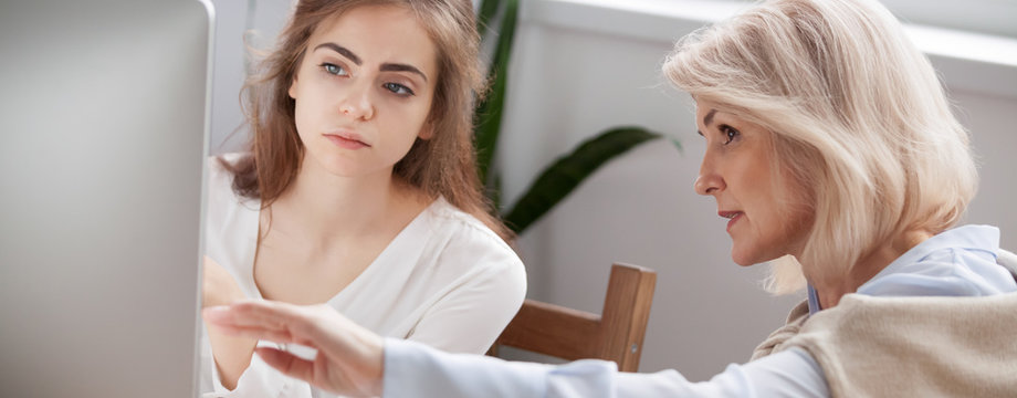 Older and younger women colleagues working on new project discuss online app point on pc screen mentor teach help intern explain corporate software, teamwork concept close up banner for website header