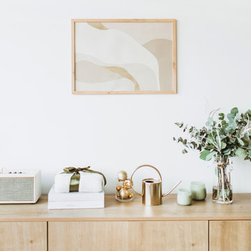 Modern interior design concept. Minimal Nordic Scandinavian style living room with photo frame on white wall, wooden table with gift box, floral bouquet with eucalyptus, candles, radio.