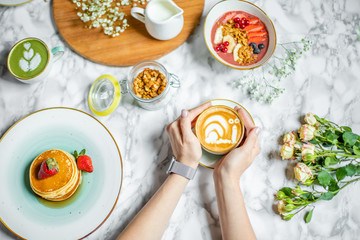 a cup of coffee with milk in hand, pancakes and cereal with fruit on the table