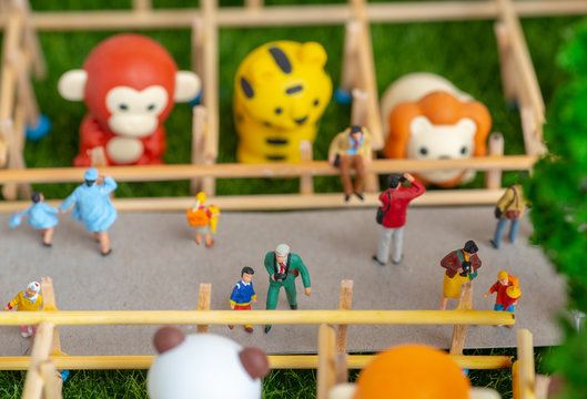 A self constructed miniature toys concept of people at the zoo - school kids, old people on benches and a photographer dangerously takes photos of his friend on a fence and a tiger. is behind him.