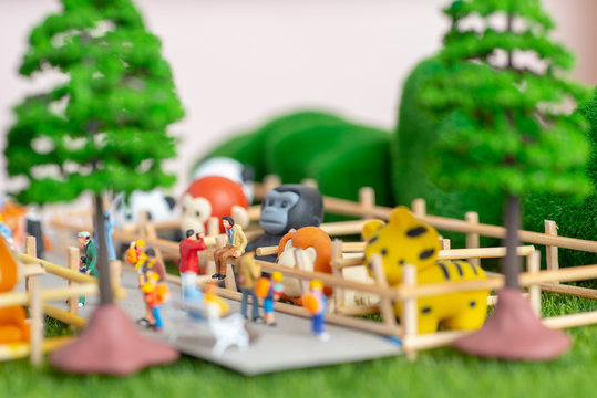 A self constructed miniature toys concept of people at the zoo - school kids, old people sitting on benches and a photographer dangerously takes photos of his friend on a fence.