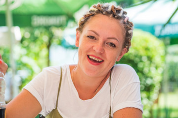Cute young girl with freckles and colorful dreadlocks is sitting at a table in an open-air cafe. Close-up portrait of a girl laughing