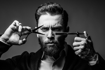 Young bearded man portrait holding Barber scissors and razor near his face standing isolated on gray background