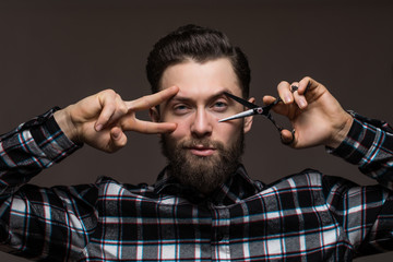 Handsome bearded young man in a plaid shirt holding a sharp professional scissors and razor isolated on dark background. Close-up