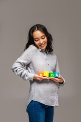 Smiling pregnant young asian woman holding alphabet cubes with kids lettering isolated on grey