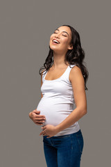 Happy pregnant asian woman with closed eyes in white t-shirt and blue jeans holding belly with hands and laughing isolated on grey