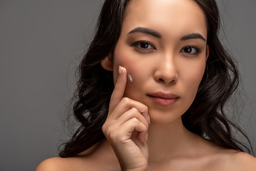Portrait of beautiful asian girl applying cosmetic cream on face and looking at camera isolated on grey