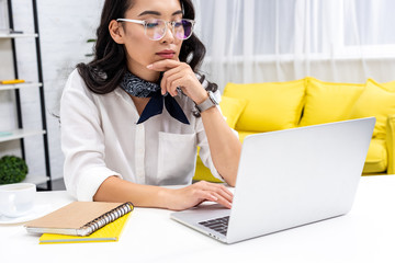 Beautiful thoughtful asian freelancer using laptop and holding hand near chin