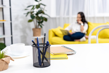 Selective focus of work desk with stationery with asian freelancer sitting on yellow sofa and using laptop on background