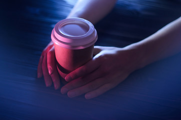 A cup of coffee on a wooden table in female hands. Paper cup Plastic cap Beautiful hands Girl bar counter light