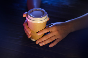 A cup of coffee on a wooden table in female hands. Paper cup Plastic cap Beautiful hands Girl bar counter light
