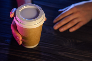 Fototapeta na wymiar A cup of coffee on a wooden table in female hands. Paper cup Plastic cap Beautiful hands Girl bar counter light