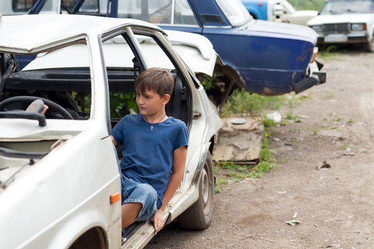 A boy, 11 years old, sits in a disassembled car in a dump of abandoned old cars on a sunny summer day.