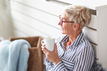 Mature good looking woman sitting in the room with mug on her hand and smile