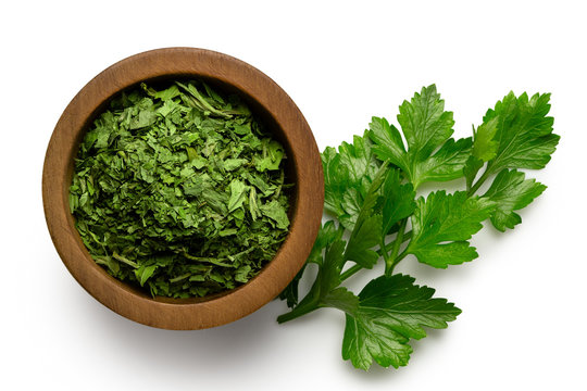 Dried chopped parsley in dark wood bowl next to fresh parsley leaves isolated on white from above.