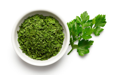 Dried chopped parsley in white ceramic bowl next to fresh parsley leaves isolated on white from...