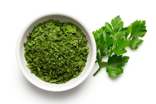 Dried chopped parsley in white ceramic bowl next to fresh parsley leaves isolated on white from above.