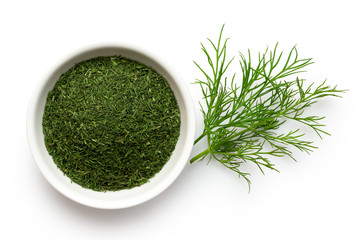 Dried chopped dill in white ceramic bowl next to fresh dill leaves isolated on white from above.
