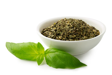 Dried chopped basil in white ceramic bowl next to fresh basil leaves isolated on white.
