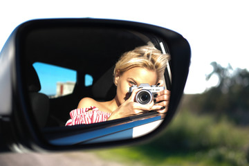 unfocused blode girl in striped red and white crop top taking photo in car mirror with vintage camera in moving car/ road trip background 