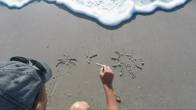 Person drawing on sand. A man paints on the sand sea sun palm trees. The sea wave washes away the painting in the sand.