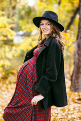 Beautiful autumn girl in the park. Lots of yellow leaves. Girl in a gray coat and red dress. Pregnant girl holding hands belly