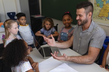 Happy male school teacher teaching schoolkid at desk in