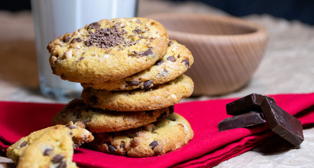 A pile of delicious and tasty chocolate chip cookies placed on the red cloth with a glass of milk in the background