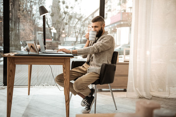Attentive good-looking bearded man in knitted cardigan sipping hot drink