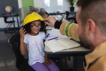 Firefighter wearing helmet to schoolgirl in classroom