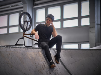 Pensive young man chilling after practicing tricks, sitting on a floor in a skatepark indoors