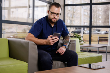 Bearded businessman dressed in a blue shirt sitting on the couch in the office, using the smartphone