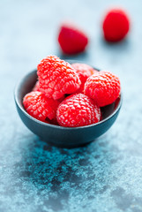 Fresh ripe raspberries in a small black bowl on a textured blue table. Macro photography.