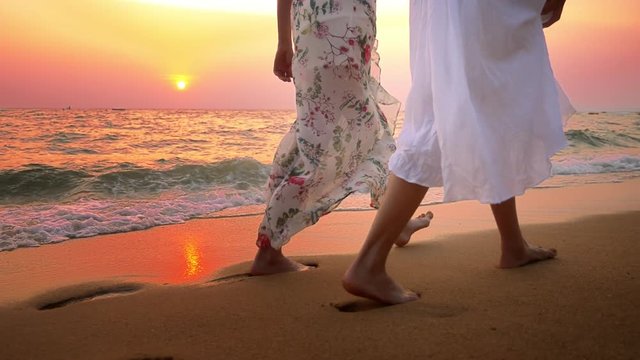 close up . bare feet on the beach. two young women in white dress walking on the beach at sunset