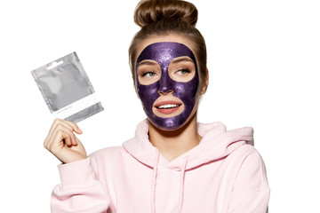 Portrait of good-looking girl cleansing skin and holding tube of mask with copy space for text. Smiling model in comfy pink sweatshirt in studio on white background