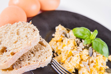 Scrambled eggs with sunflower and pumpkin seeds, some fresh eggs and wholemeal bread, eaten with a fork, decorated with basil, on a white background