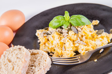 Scrambled eggs with sunflower and pumpkin seeds, some fresh eggs and wholemeal bread, eaten with a fork, decorated with basil, on a white background
