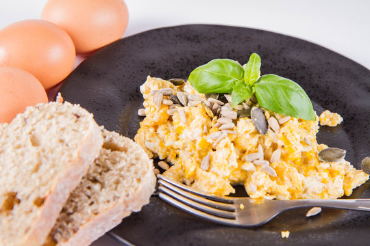 Scrambled eggs with sunflower and pumpkin seeds, some fresh eggs and wholemeal bread, eaten with a fork, decorated with basil, on a white background