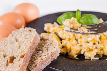 Scrambled eggs with sunflower and pumpkin seeds, some fresh eggs and wholemeal bread, eaten with a fork, decorated with basil, on a white background