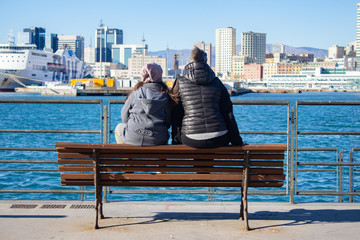 Mother and daughter sitting on a bench, watching the sea of the port of Genoa 2
