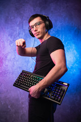 Young handsome gamer wearing headset with microphone, holding a keyboard and pointing finger at camera on neon colored background - obrazy, fototapety, plakaty