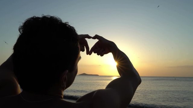 Young man with curly hair making heart with his hands at amazing sunset on the beach. Silhouette of dreamer. Art inspiration and beautiful light.